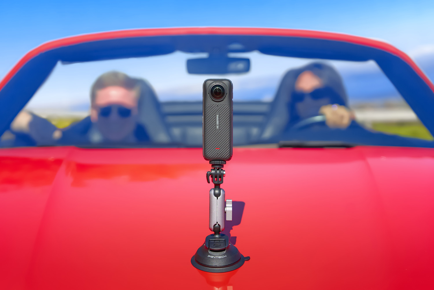 Камера обзора 360 градусов, установленная на капоте красного автомобиля, на заднем плане видны два размытых человека: один за рулем, другой на пассажирском сиденье, оба в солнцезащитных очках.