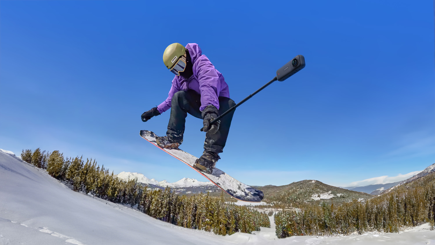 Сноубордист находится в воздухе во время прыжка, в фиолетовой куртке и зимних штанах, в шлеме и очках, с 360-градусной камерой, прикрепленной к концу палки для селфи, на фоне заснеженных гор.